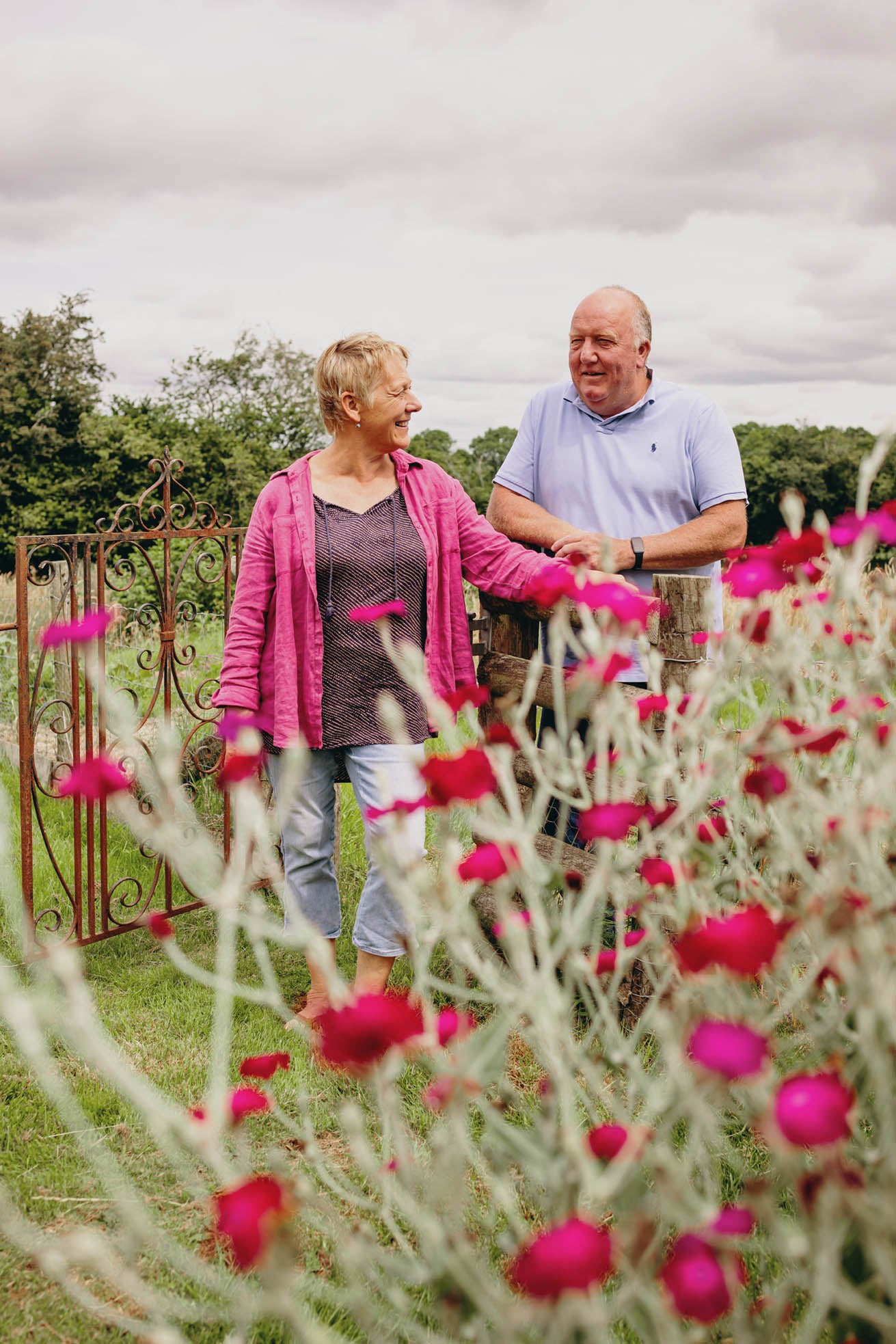 An elderly couple smiling and standing in a garden. The woman in a pink jacket laughs with the man in a light blue shirt. They are near an ornate metal gate, surrounded by vibrant pink and red flowers under a cloudy sky.