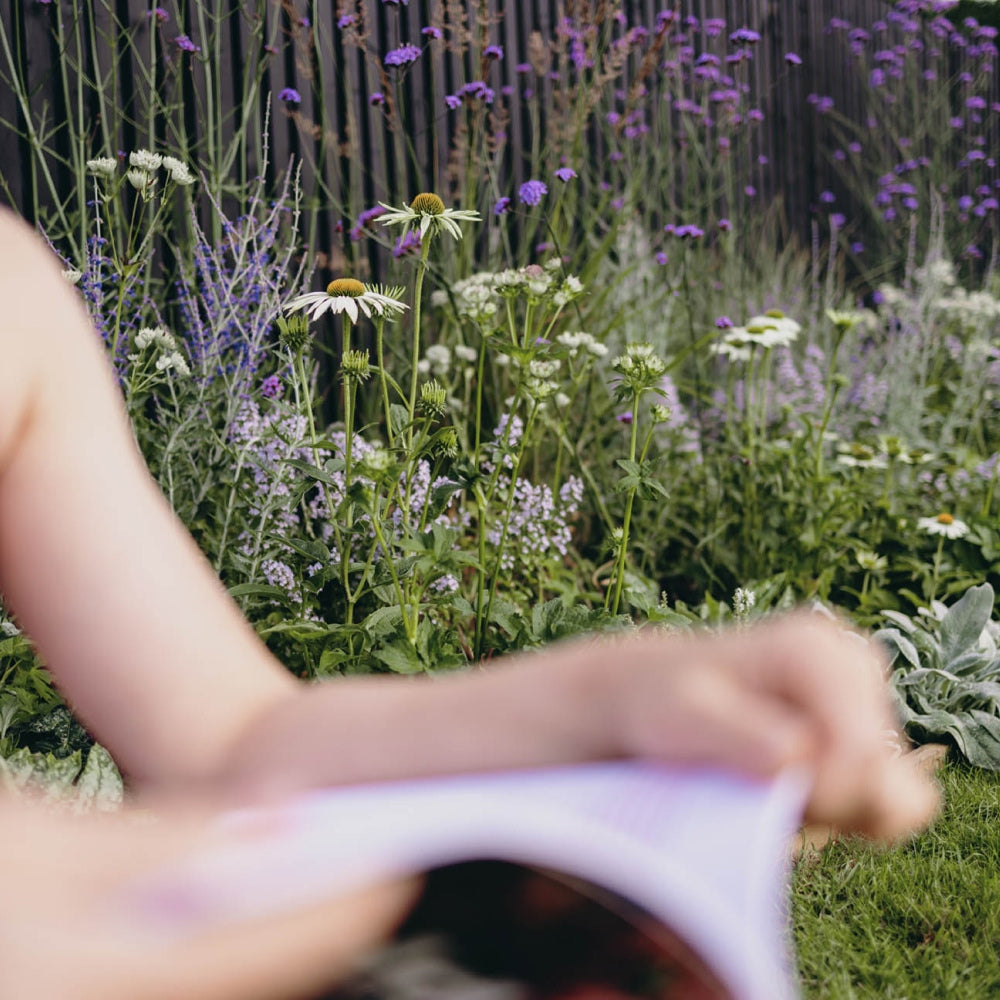 A person holding a magazine is sitting in a garden. The foreground is blurred, focusing on a lush garden with white and purple flowers and green foliage. A wooden fence is in the background.