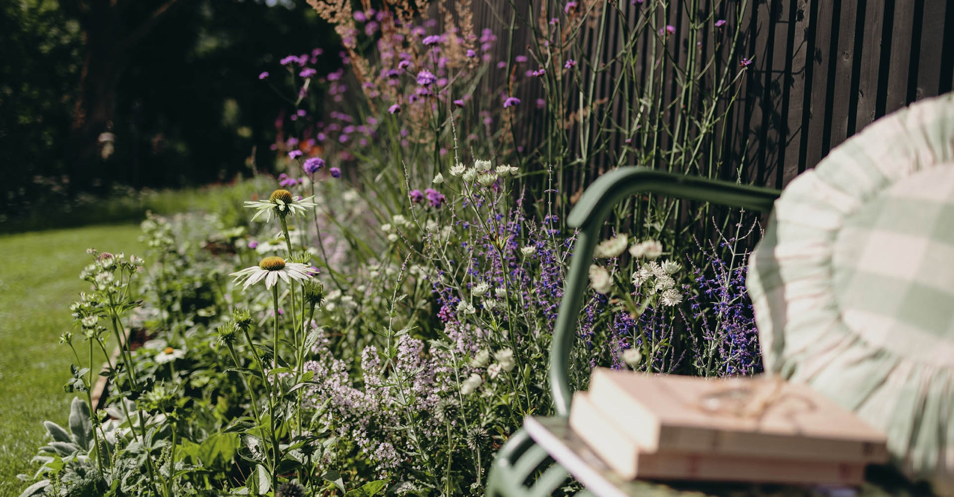 A garden scene with purple and white flowers growing along a black fence. A green chair with a plaid cushion sits in the foreground, holding a stack of books. The grass is lush and the sunlight creates dappled shadows on the ground.